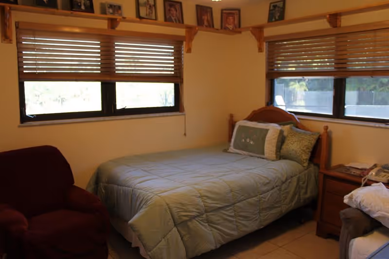 A cozy bedroom with a single bed covered in a light green quilt and multiple pillows. There are two windows with wooden blinds on adjacent walls. A wooden nightstand with a telephone and some papers is next to the bed. A maroon armchair is positioned near the bed. Above the windows, a wooden shelf holds framed photographs.