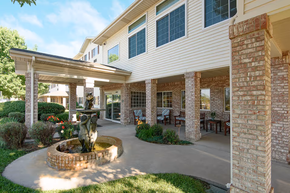 Covered entrance of a senior living building with brick columns, a small fountain, and outdoor seating.