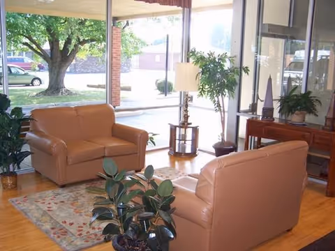 A bright living room area with two tan leather sofas facing each other on a patterned rug. There are several potted plants around the room, a small round table with a lamp between the sofas, and large windows letting in natural light with a view of trees and parked cars outside.