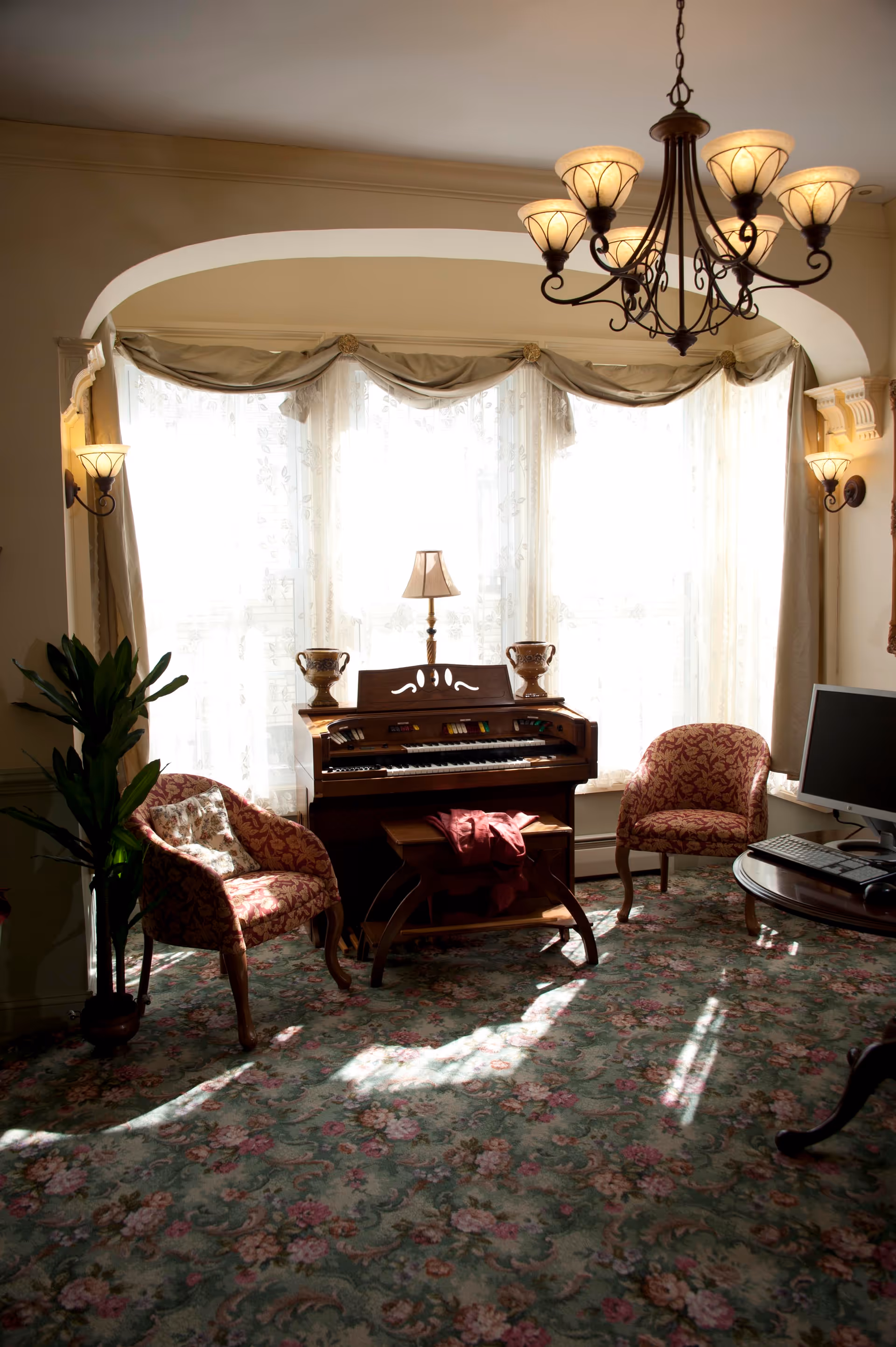 A cozy living room area with floral carpet, two upholstered armchairs with patterned fabric, a small wooden organ with a lamp and decorative items on top, a round wooden table with a computer monitor and keyboard, and a large window with sheer curtains letting in natural light. A chandelier and wall sconces provide additional lighting.