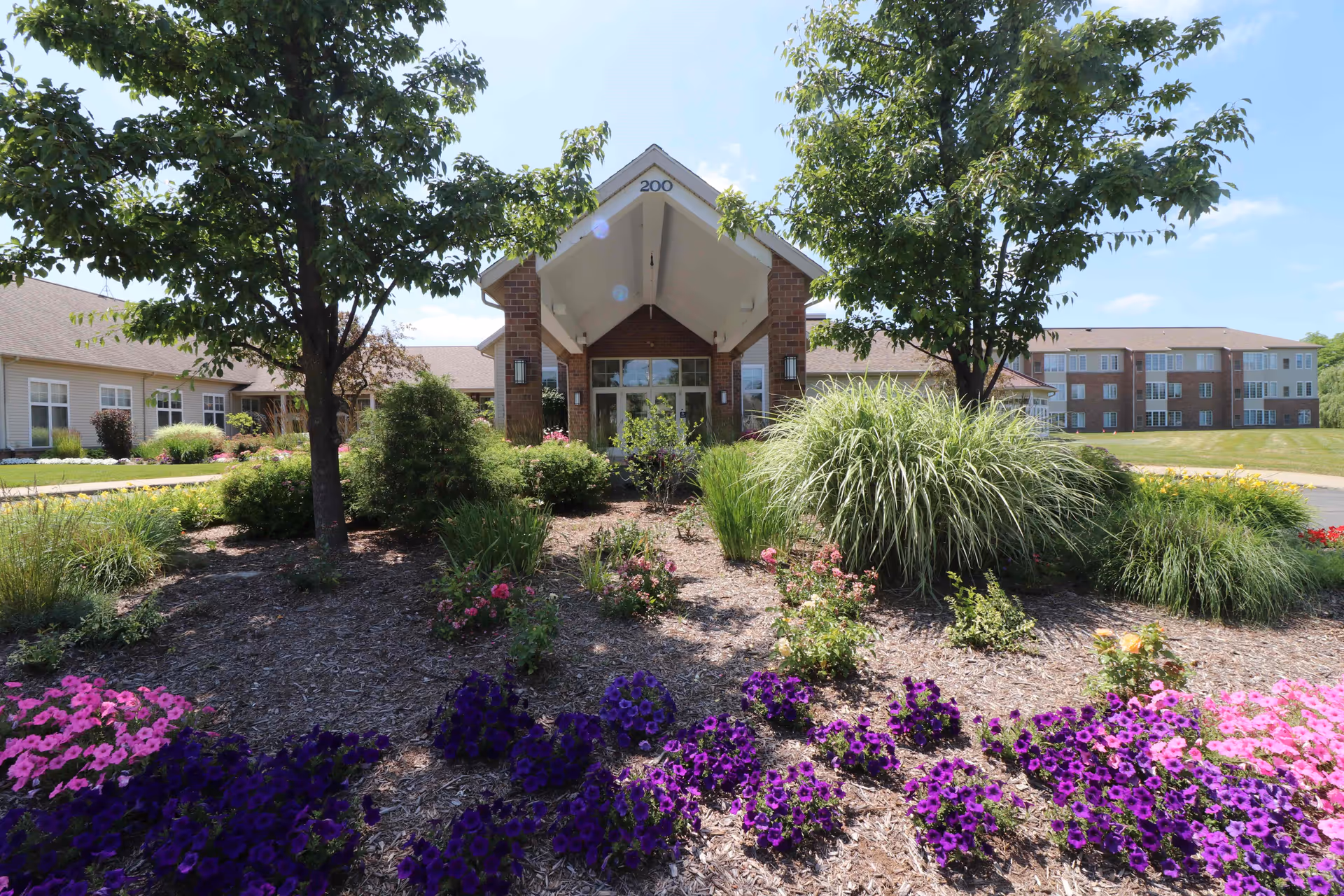 Front entrance of a brick senior living building with a porte-cochère, surrounded by trees and colorful flower beds.