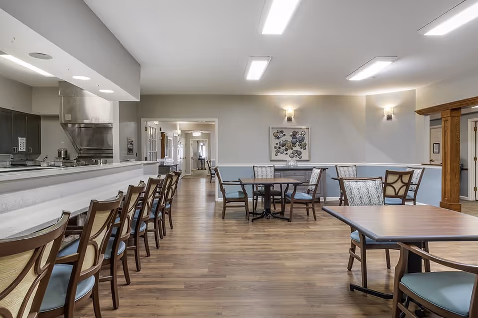 Interior view of a dining room at Brookdale Dublin, featuring tables and chairs, a kitchen area, and light-colored walls.
