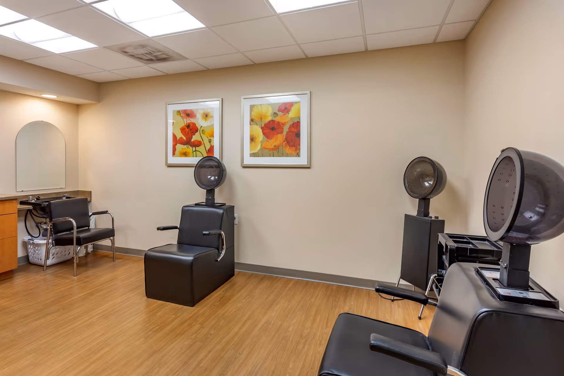 Interior of a senior community hair salon with three black salon chairs, two with hair dryers attached. There is a small sink with a chair next to it, wooden flooring, beige walls, and two framed floral paintings on the wall.