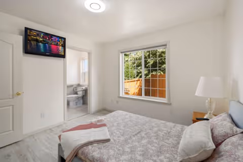 A bedroom with a bed covered in a patterned bedspread and pillows, a bedside table with a lamp, a window showing a wooden fence and greenery outside, and an open door leading to a bathroom with a toilet and sink. A colorful picture is mounted on the wall above the bathroom door.