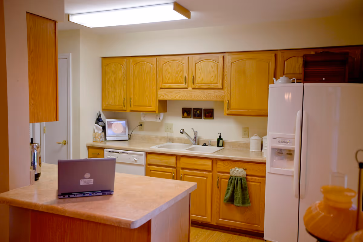 A kitchen with wooden cabinets, a double sink, a white refrigerator with an ice and water dispenser, a dishwasher, and a countertop island with a laptop on it. There is a small digital photo frame and a tissue box on the counter near the sink.