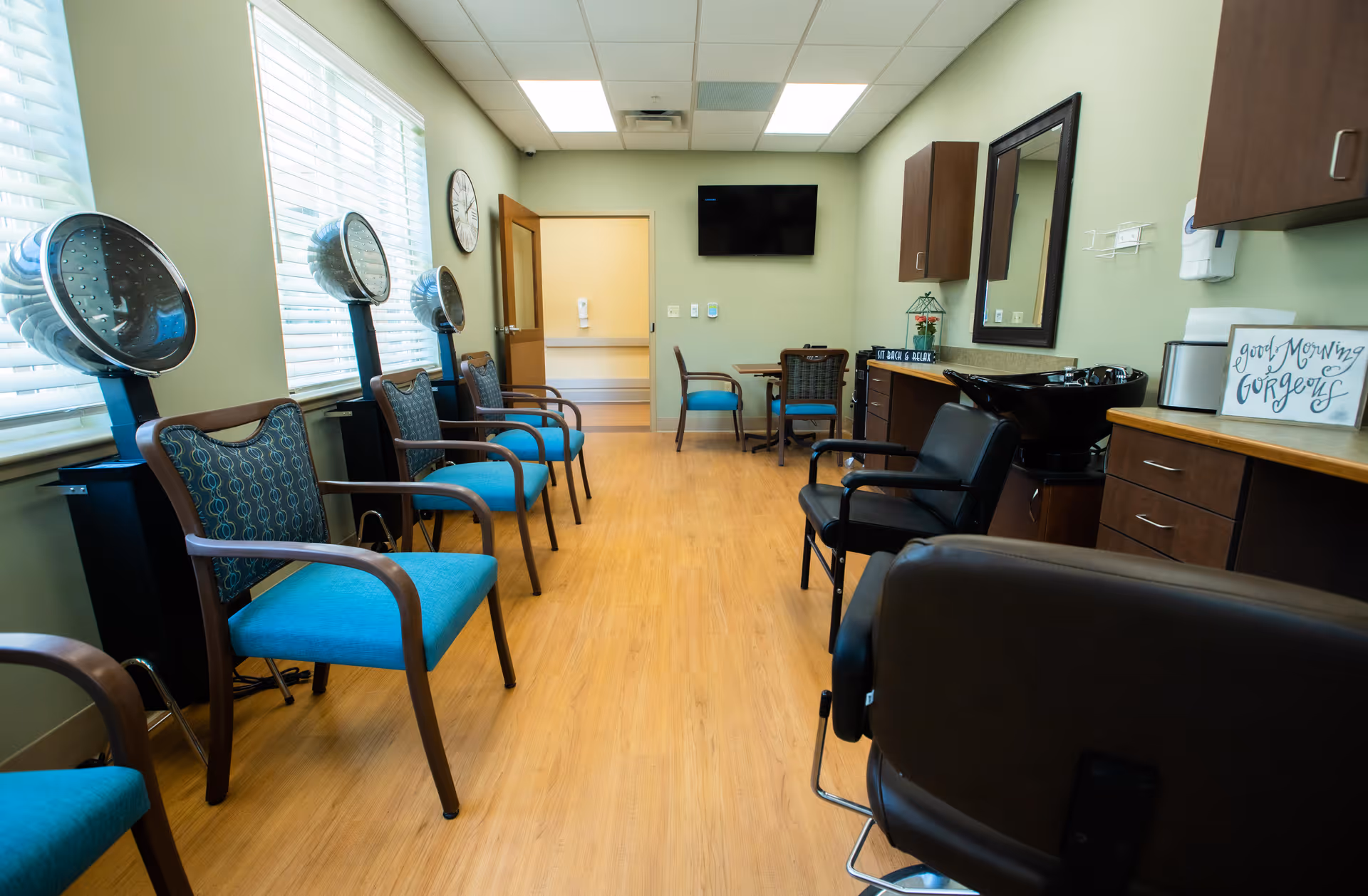 Interior view of a hair salon area in a senior living facility with several blue cushioned chairs lined up along the left wall under hair dryers. On the right side, there are black salon chairs, a sink for washing hair, cabinets, and a mirror. The room has light green walls, wood flooring, a wall-mounted TV, and a small table with chairs in the back.