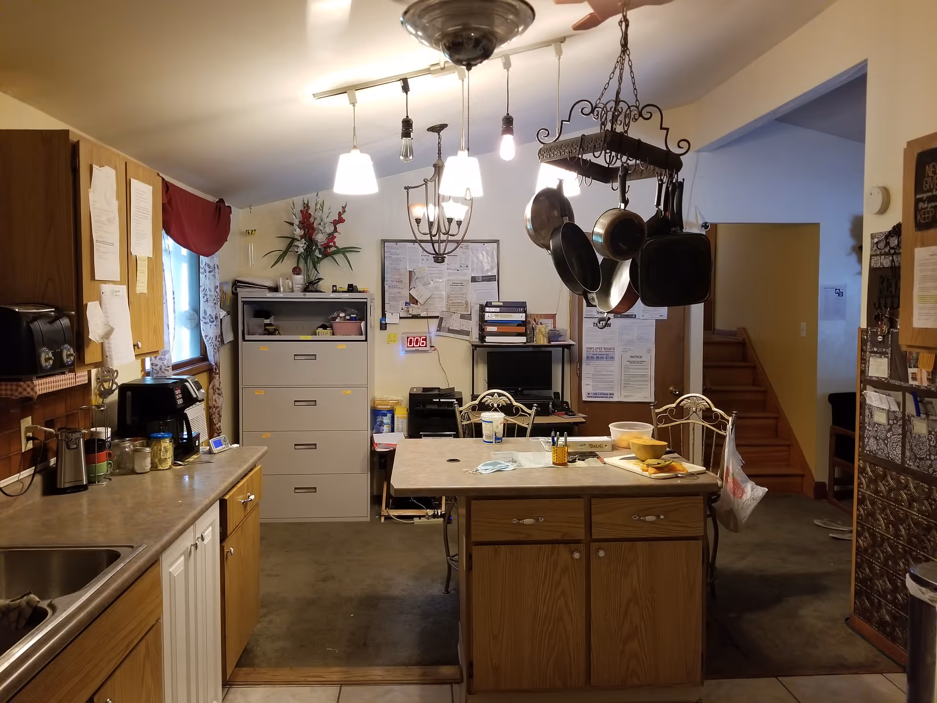 Interior view of a kitchen area with wooden cabinets, a countertop with various kitchen items, a hanging pot rack with pots and pans, a filing cabinet, and a table with chairs. There are papers and office supplies on the table, a bulletin board on the wall, and a staircase leading to another room.