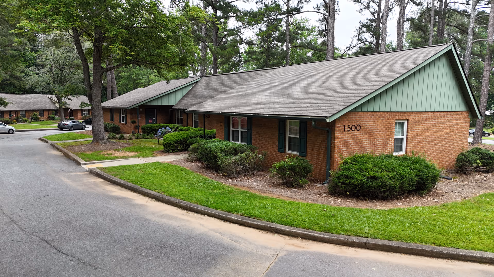 Exterior view of a single-story brick building with green trim and a gray roof, surrounded by grass, bushes, and trees. The building has the number 1500 on the side. A paved road curves around the building, and other similar buildings and parked cars are visible in the background.