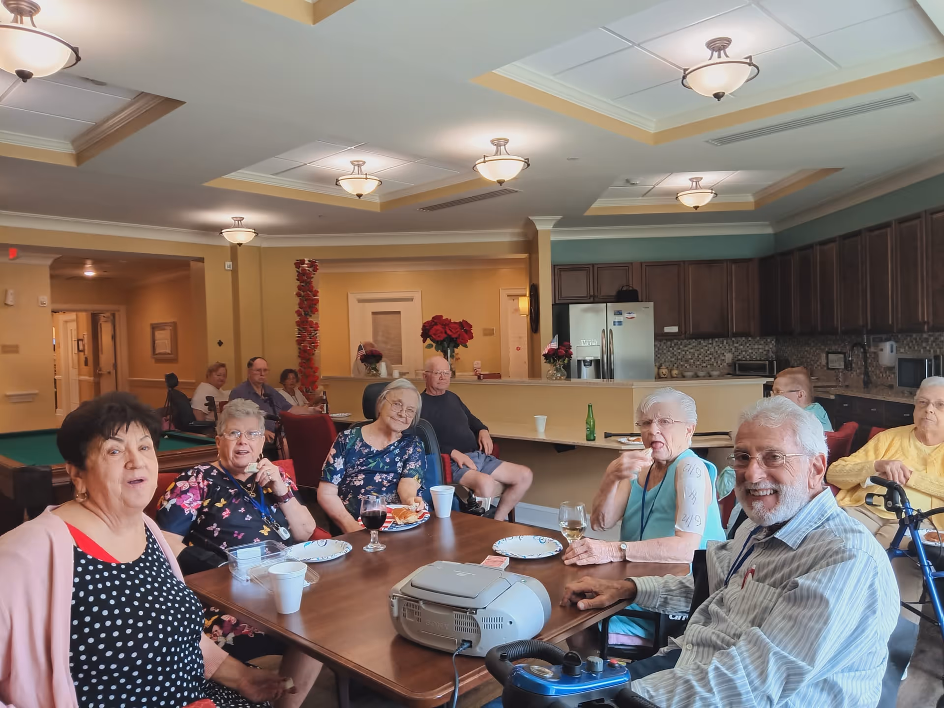 A group of elderly residents seated around a dining table in a senior living community common room, eating and socializing.