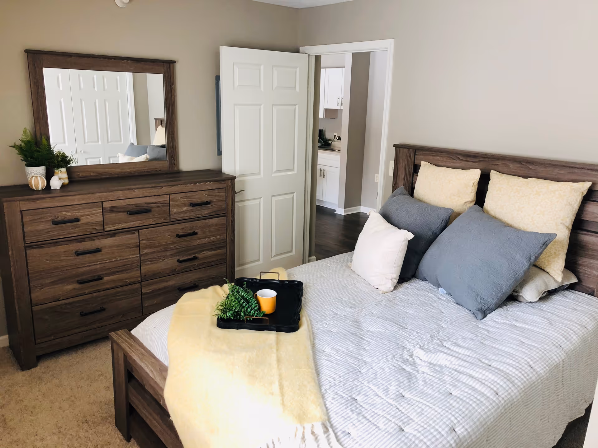 A bedroom with a wooden bed frame and a bed made with white and gray bedding and multiple pillows. A yellow blanket and a black tray with a cup and greenery are placed on the bed. There is a wooden dresser with a large mirror above it, decorated with small plants and ornaments. The door to the room is open, showing a glimpse of a kitchen area with white cabinets.