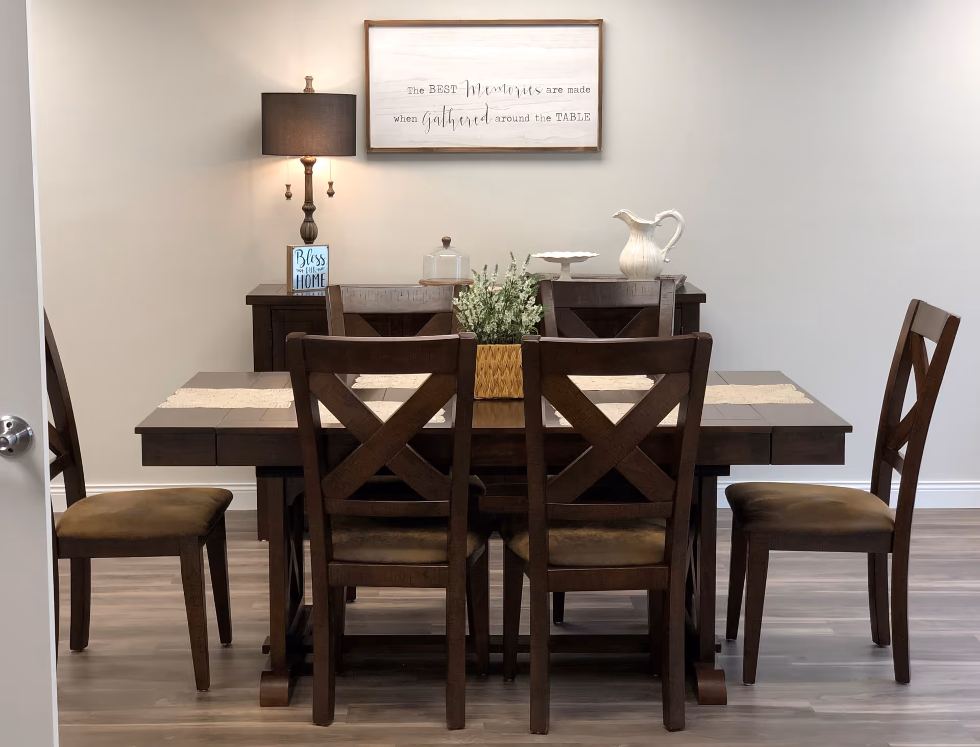 Wooden dining table with six chairs, a floral centerpiece, and decorative items against a gray wall with a framed sign and lamp.