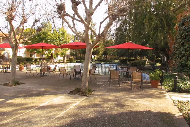 Outdoor courtyard with patio tables, red umbrellas, and a pool surrounded by trees and walkways.