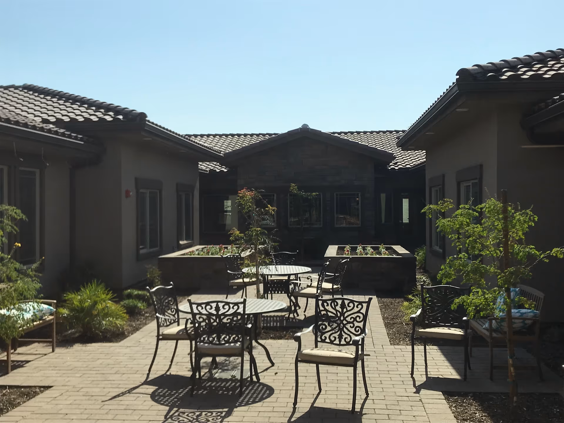 Outdoor courtyard area with metal tables and chairs arranged on a paved patio, surrounded by low garden beds with plants and small trees, flanked by single-story buildings with tiled roofs under a clear blue sky.