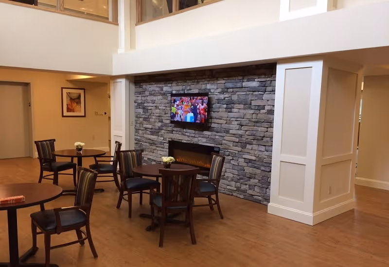 Communal lounge with round tables and chairs facing a stone accent wall with a mounted TV and electric fireplace.
