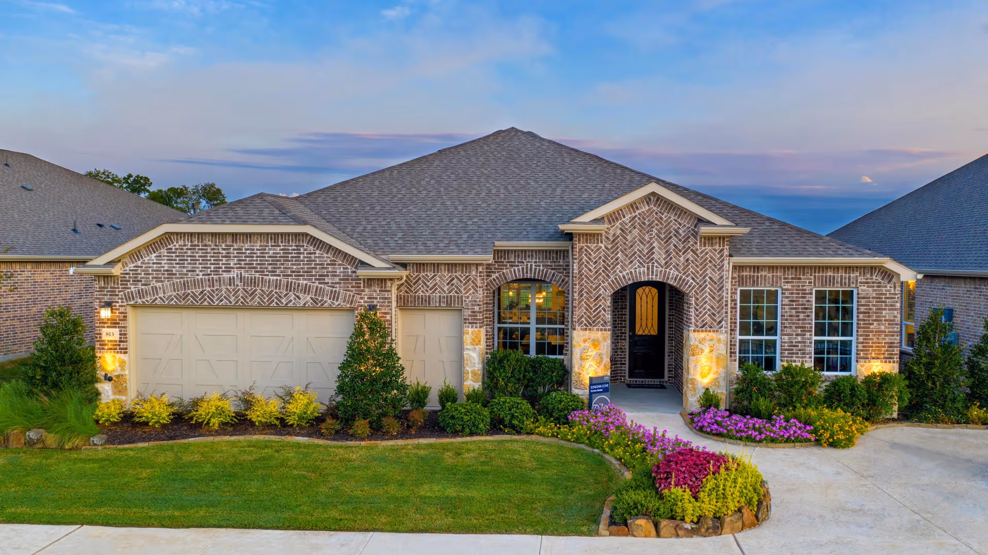 Front exterior view of a single-story brick house with a two-car garage, well-maintained lawn, and colorful flower beds under a partly cloudy sky at dusk.