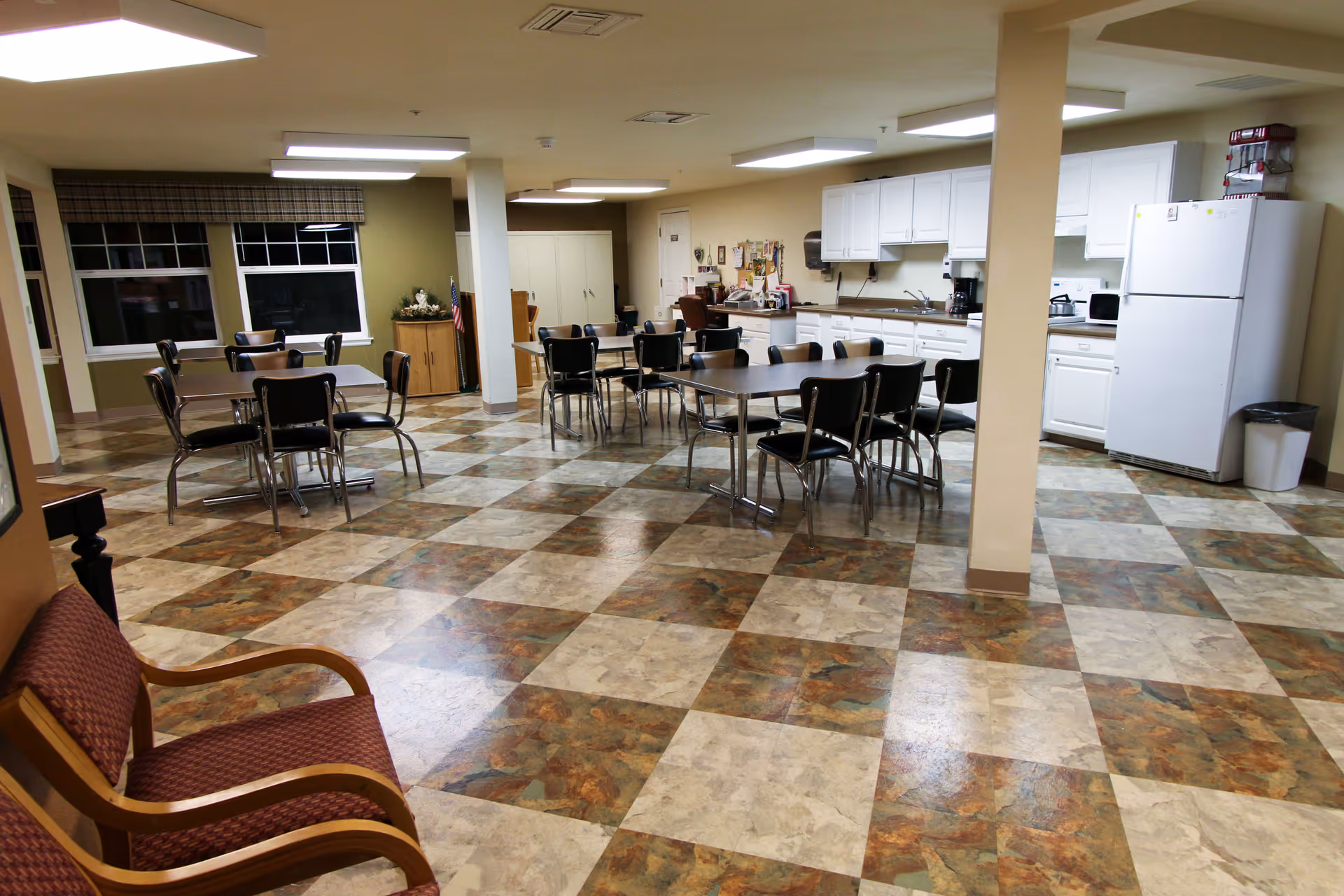 A spacious common area in a senior living facility featuring a kitchen with white cabinets, a refrigerator, and a microwave. Several tables with black chairs are arranged in the room, and there is a seating area with wooden armchairs with red cushions. The floor has a checkered pattern with brown and beige tiles, and the room is well-lit with ceiling lights.