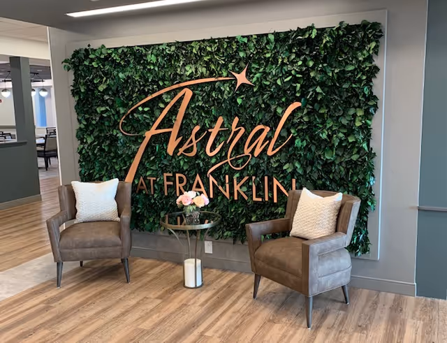 Two brown armchairs with white pillows are placed on either side of a small round glass table with a flower arrangement. Behind them is a green leafy wall with the words 'Astral at Franklin' in large decorative letters.