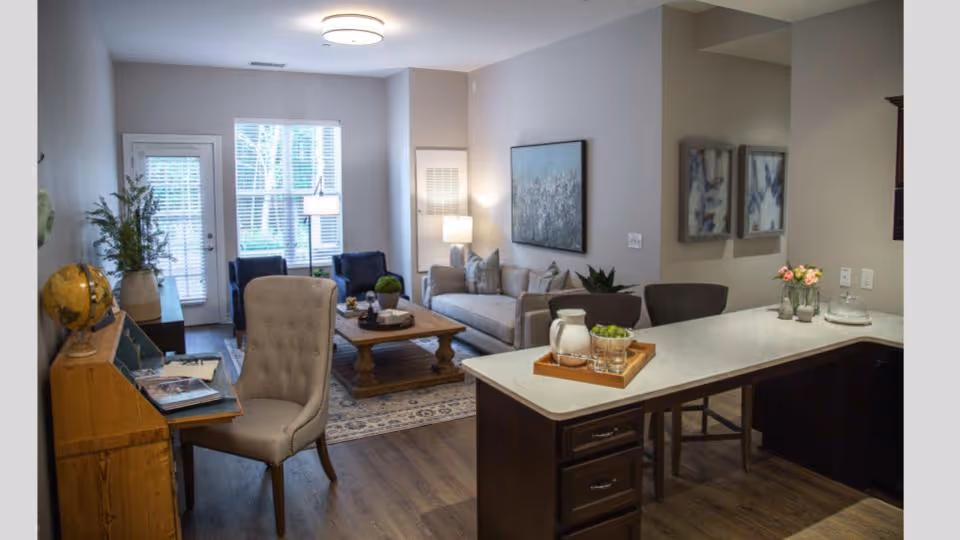 A cozy living room and kitchen area in a senior living facility. The living room features a beige sofa, two dark blue armchairs, a wooden coffee table, and a floor lamp near a window with blinds. The kitchen has a white countertop with a tray holding a pitcher, glasses, and a bowl of green grapes. There is a wooden desk with a globe and books, and a beige upholstered chair in the foreground. The walls are painted light gray, and there are framed artworks on the walls.