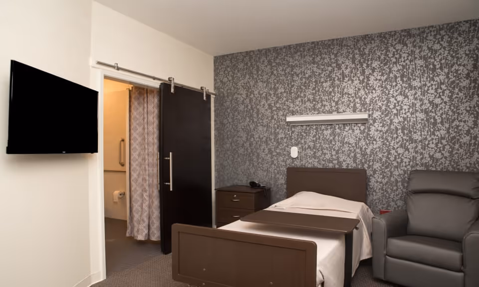 A senior living facility bedroom with a single bed, a bedside table with a phone, a gray armchair, and a wall-mounted TV. The room has a patterned gray accent wall and a sliding barn door leading to a bathroom with a shower curtain visible.