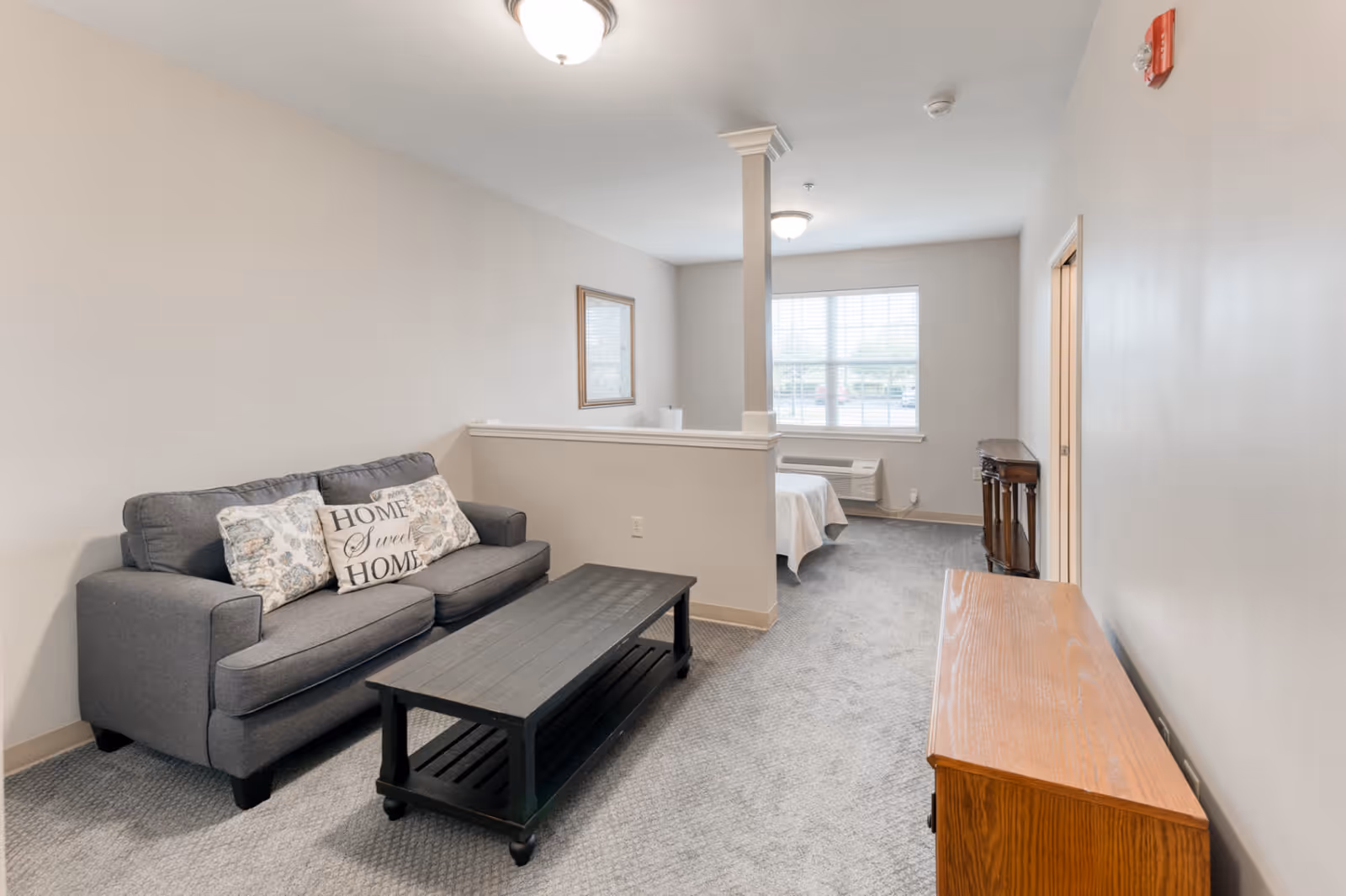 A cozy living area with a gray sofa adorned with decorative pillows, including one that says 'Home Sweet Home.' In front of the sofa is a black coffee table. The room has light-colored walls and carpeted flooring. A half wall separates the living area from a bedroom space with a bed near a window letting in natural light. There is also a wooden cabinet and a small table against the walls.