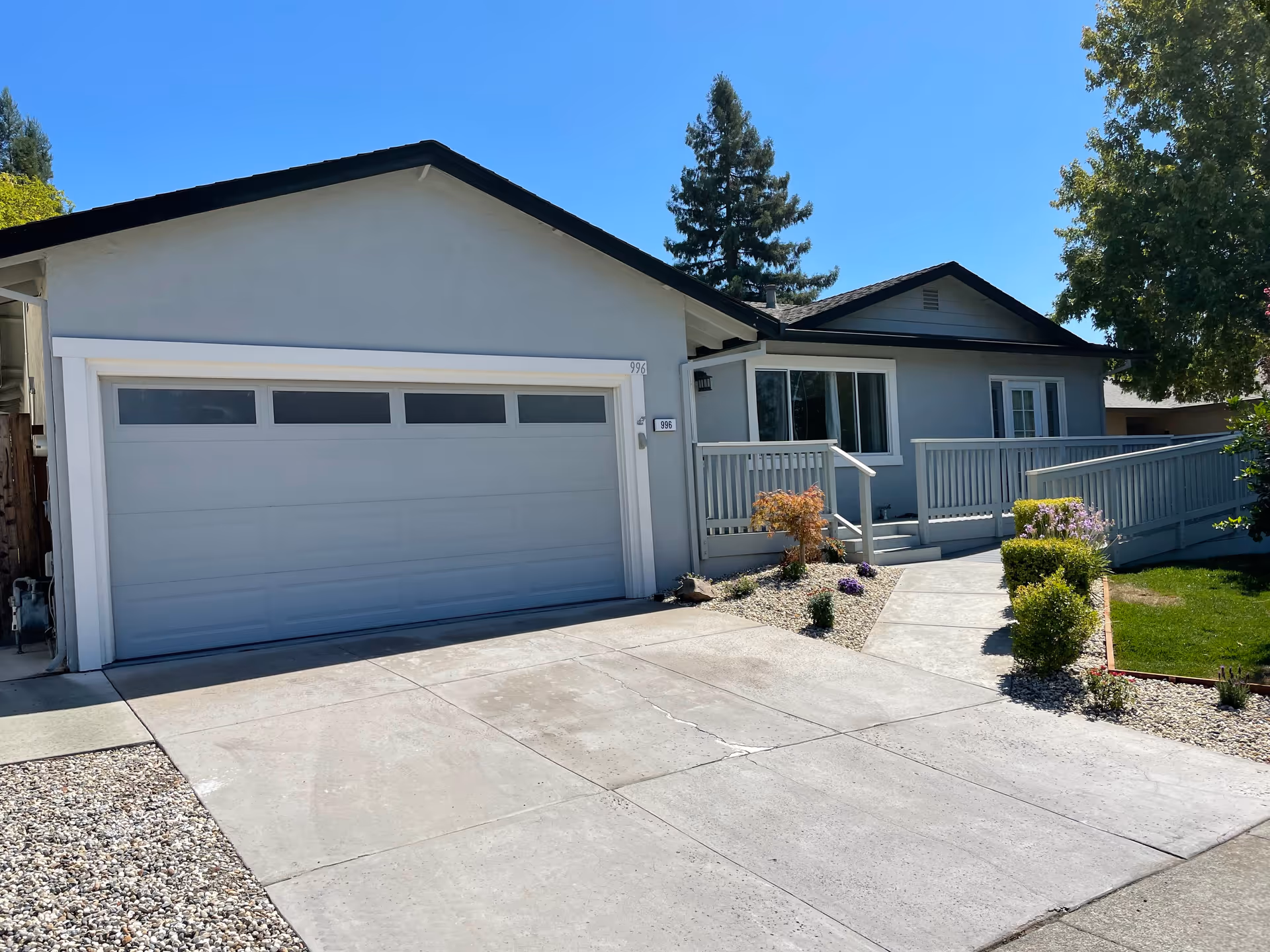 Exterior view of a single-story residential building with a two-car garage, a concrete driveway, a small garden with shrubs and flowers, and a wheelchair-accessible ramp leading to the front entrance. The sky is clear and blue.