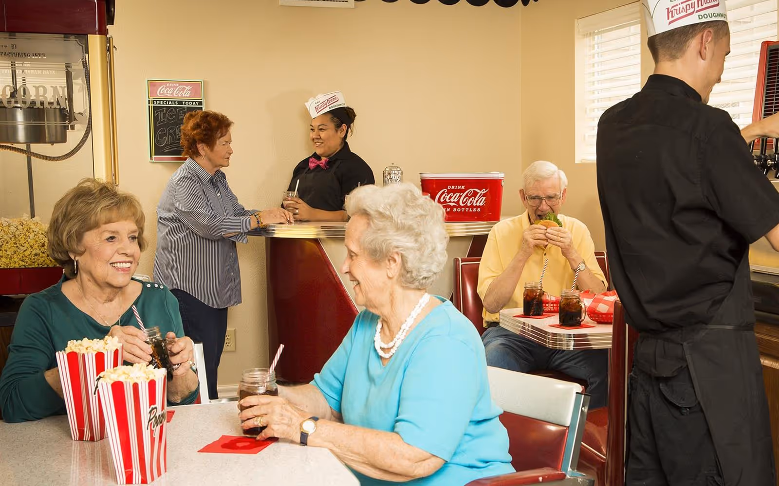 A retro-style diner scene in a senior living facility with elderly people enjoying snacks and drinks. Two elderly women sit at a table with popcorn and drinks, smiling and chatting. An elderly man is seated in a booth eating a sandwich with two drinks on the table. A server wearing a Krispy Kreme hat is pouring a drink, while another staff member in a similar hat talks to a guest at the counter. The setting includes vintage decor such as a popcorn machine and Coca-Cola cooler.