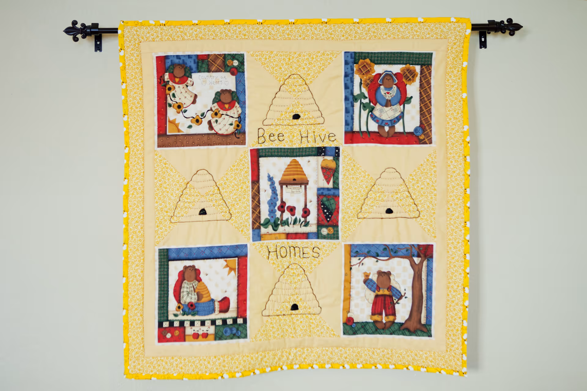 A decorative quilt hanging on a wall with a yellow border and various patchwork designs including bears, beehives, flowers, and strawberries. The quilt has the words 'Bee Hive Homes' stitched in the center.
