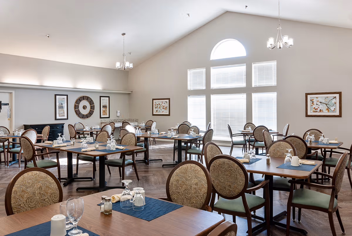 Bright dining room with multiple set tables and upholstered chairs under a vaulted ceiling and large windows.