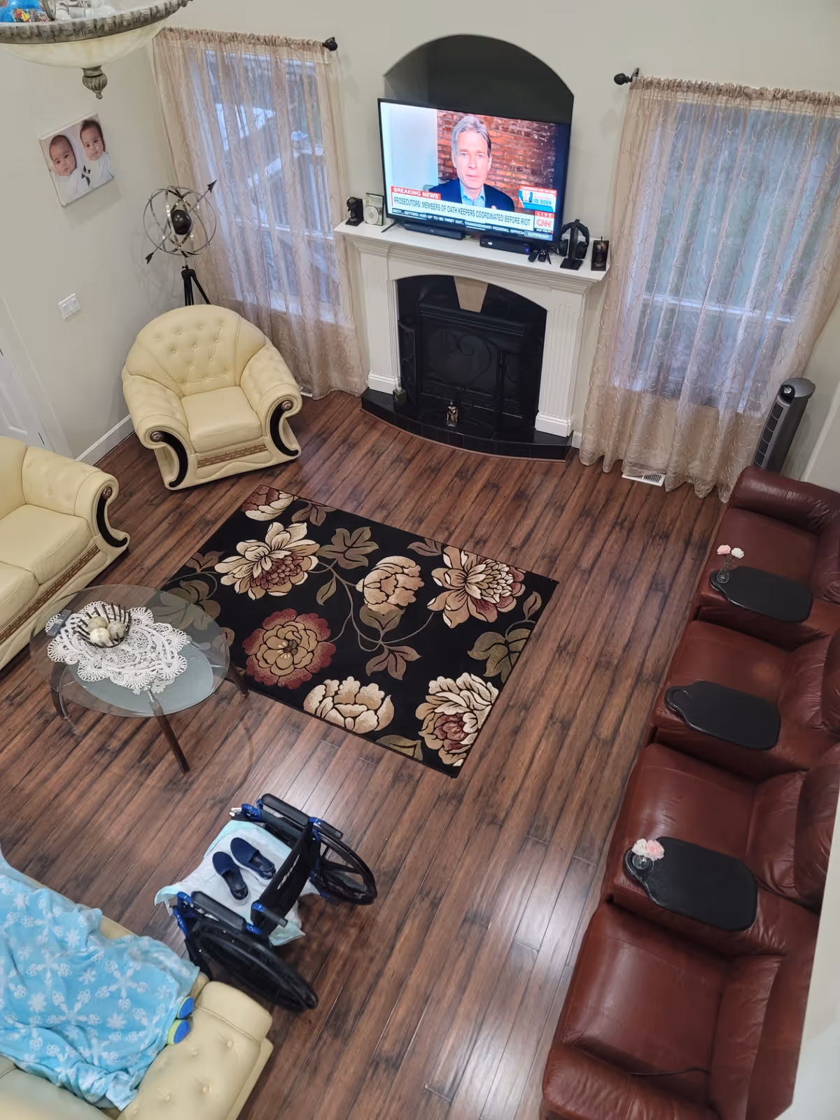 A cozy living room with wooden flooring, a floral area rug, and a fireplace with a TV mounted above it. The room has cream-colored armchairs and a sofa, a maroon leather recliner sofa with black trays, sheer curtains on the windows, and a glass coffee table with a doily and decorative items. A wheelchair is positioned near one of the armchairs.