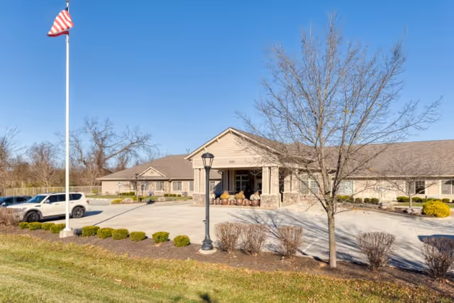 Exterior view of Cedar Creek of Bloomington Memory Care facility showing a single-story building with a covered entrance, a flagpole with an American flag, a parking area with a white SUV, and landscaped bushes and trees under a clear blue sky.