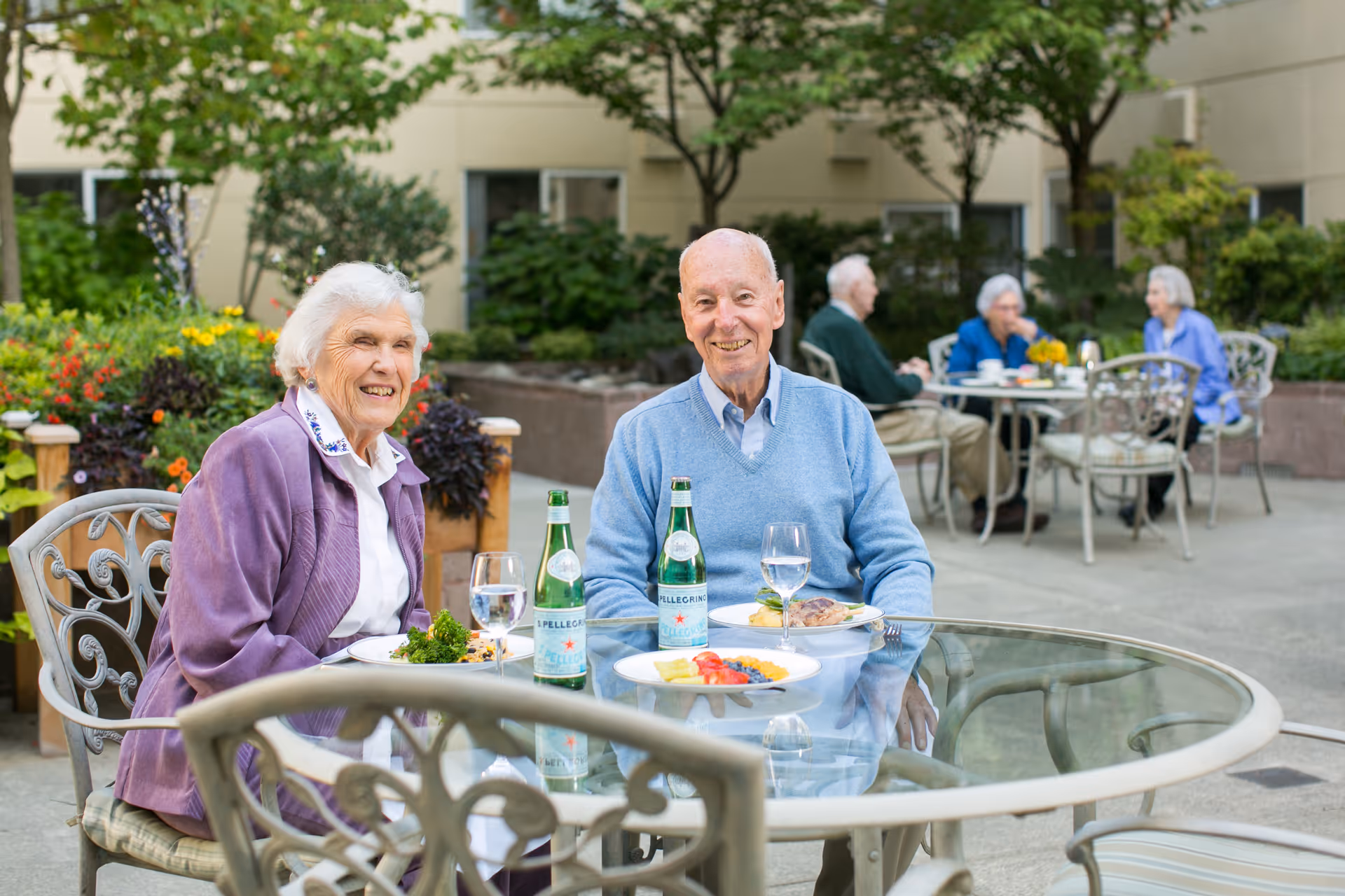 Two elderly people, a woman and a man, sitting at a glass outdoor table with plates of food and bottles of sparkling water, smiling at the camera. In the background, three other elderly people are seated at another table in a garden courtyard with greenery and flowers.