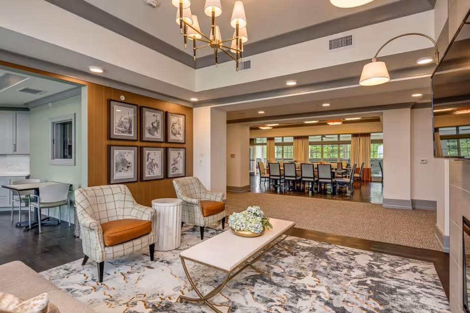 Bright communal living area with upholstered chairs, a coffee table, and a dining table visible in the background.
