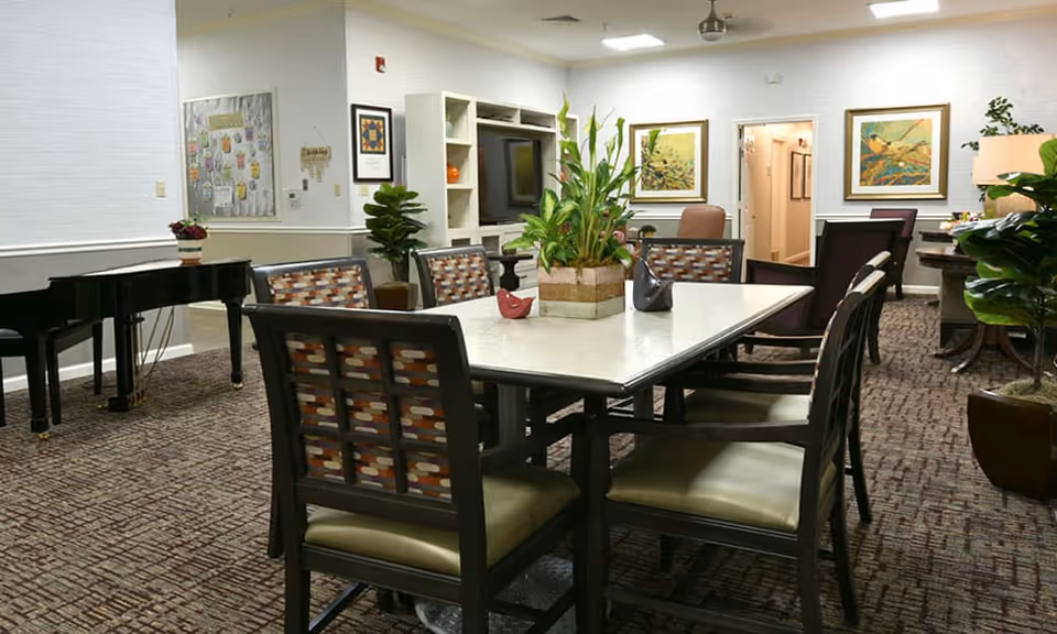 A well-lit common area with a rectangular table surrounded by eight chairs, a decorative plant centerpiece on the table, a black grand piano on the left side, framed artwork on the walls, and a carpeted floor. There are also plants and a lamp in the room, with an open doorway leading to a hallway in the background.