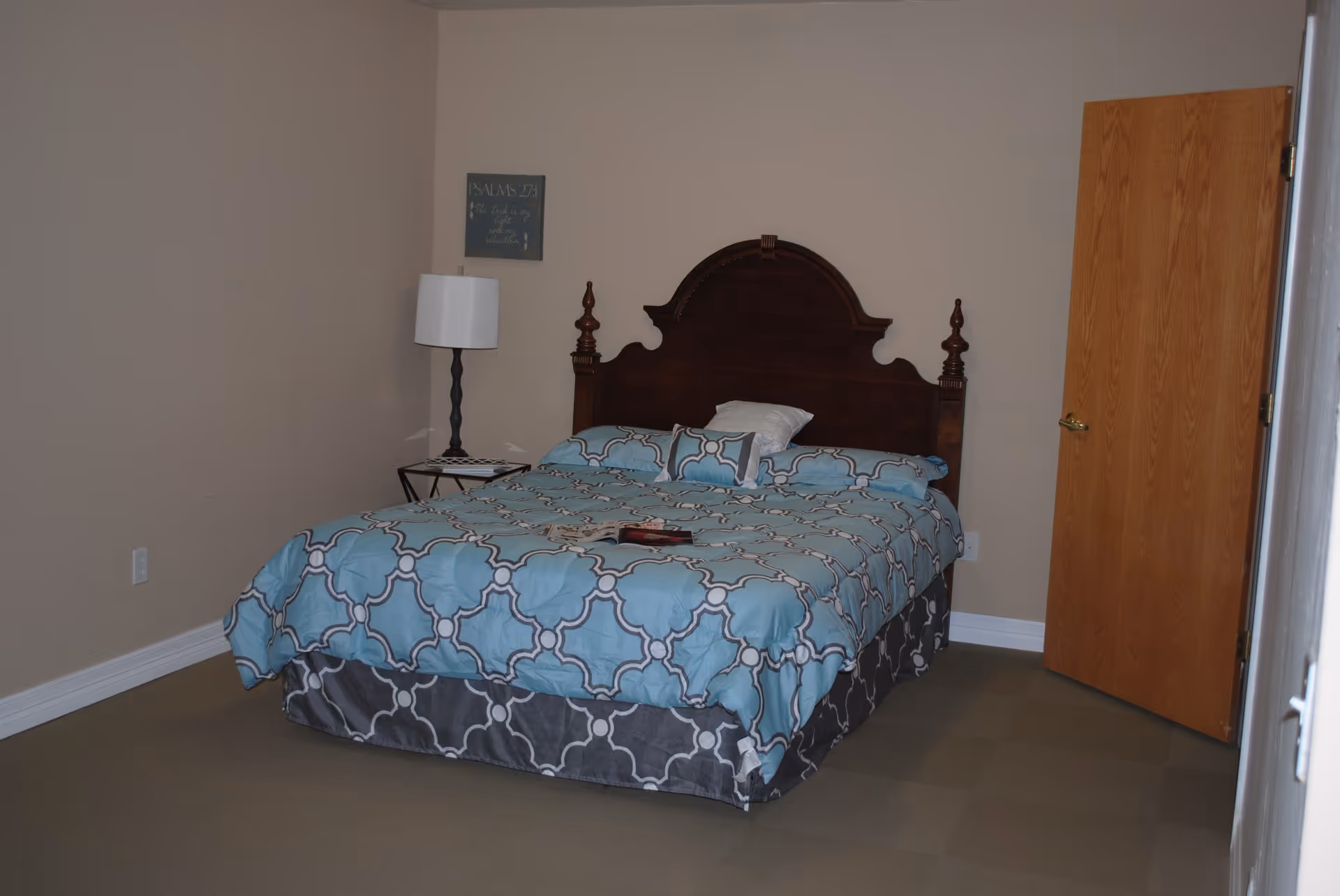 Bedroom with a wooden headboard and a bed covered in blue patterned bedding, a bedside table with lamp, and a partially open wooden door.
