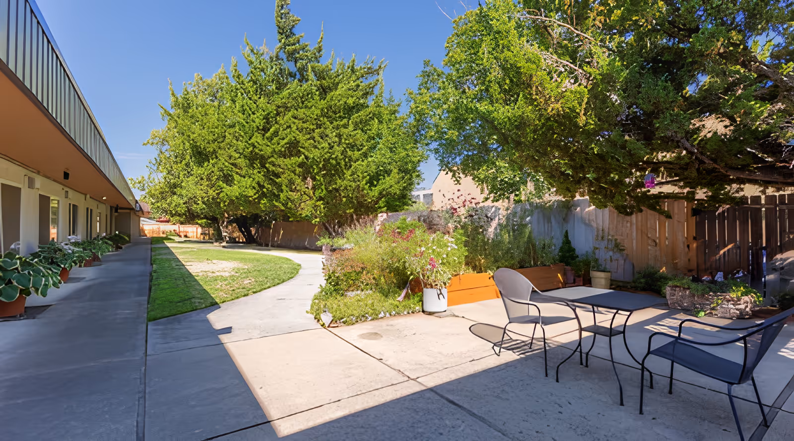 Outdoor patio area at Hearthstone Nursing & Rehabilitation Center featuring a concrete walkway, potted plants, garden beds, trees providing shade, and a metal table with chairs under a tree on a sunny day.