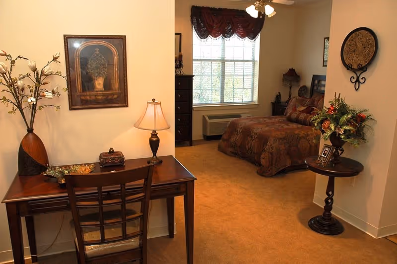 A cozy assisted-living bedroom with a wooden desk and chair in the foreground, decorative lamp and vase, and a bed by a window in the background.