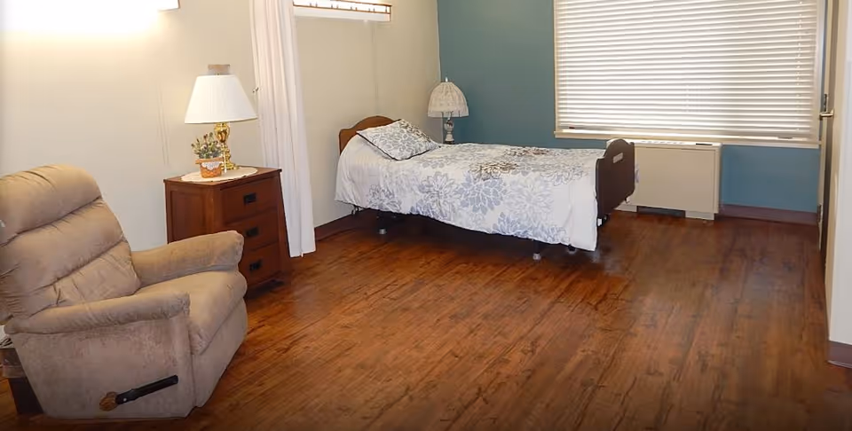 A simple nursing facility bedroom with a single bed, recliner, bedside table and lamp, wood-look floor and a window with blinds.