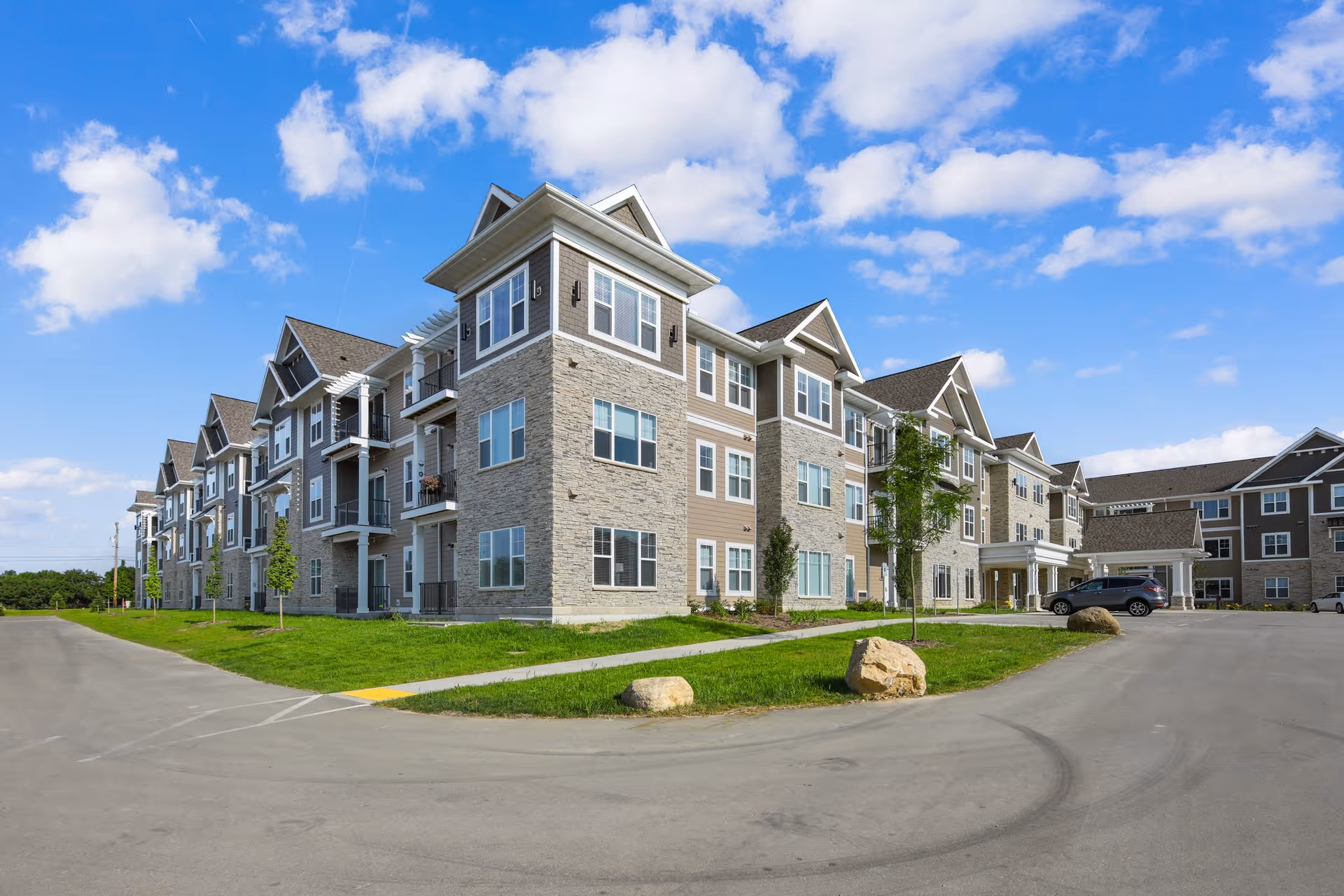 Large three-story residential building with stone and siding, a parking area and landscaped lawn under a blue sky.