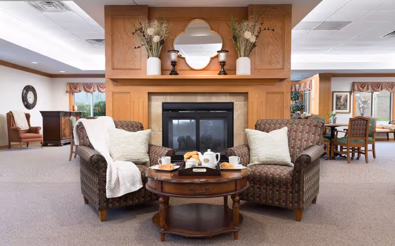 A cozy living room area in a senior living facility featuring two patterned armchairs with light-colored pillows and a white throw blanket. Between the chairs is a wooden coffee table set with a white teapot, cups, plates with pastries, and a small tray. Behind the chairs is a fireplace with a wooden mantel decorated with a mirror, two white vases with dried flowers, and two candle holders. The room has carpeted floors, large windows with valances, and additional seating and tables in the background.
