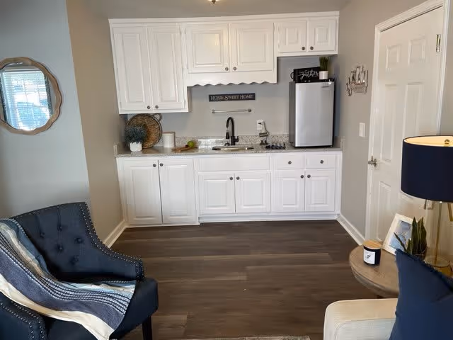 A small kitchenette area with white cabinets, a granite countertop, a black faucet, and a mini refrigerator. There is a decorative sign that says 'HOME SWEET HOME' on the wall above the sink. To the left, there is a dark blue armchair with a striped throw blanket draped over it. To the right, part of a beige sofa and a round side table with a lamp, a framed photo, and a small plant are visible. The floor is dark wood, and a round mirror hangs on the left wall.