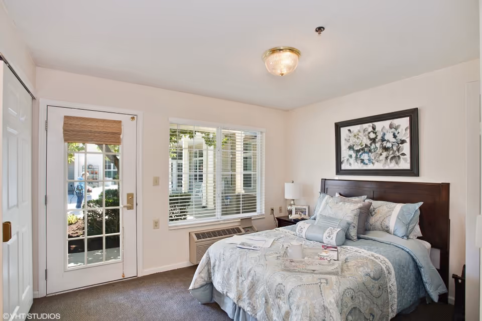 A bright bedroom with a large window and a glass door leading outside. The room features a neatly made bed with patterned bedding and multiple pillows, a bedside table with a lamp and framed photo, and a framed floral painting above the headboard. The floor is carpeted and there is an air conditioning unit below the window.