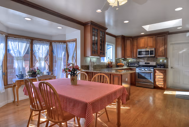 Bright open kitchen with wooden cabinets, stainless steel appliances, an island, and a dining table with a red checkered tablecloth by bay windows.