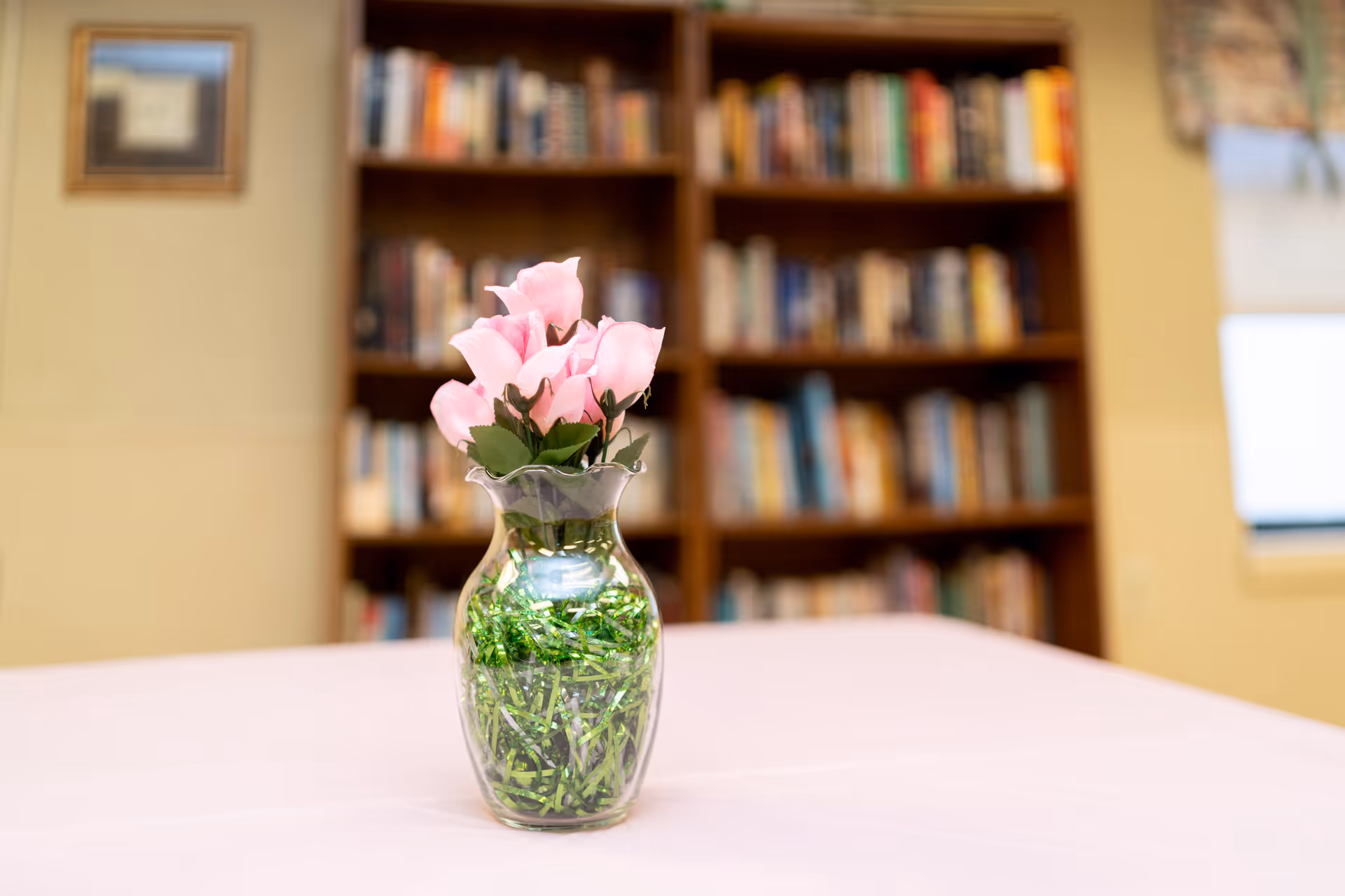 A clear glass vase with pink artificial flowers and green decorative filler sits on a table covered with a light pink tablecloth. In the background, there is a wooden bookshelf filled with books and a framed picture on the wall.