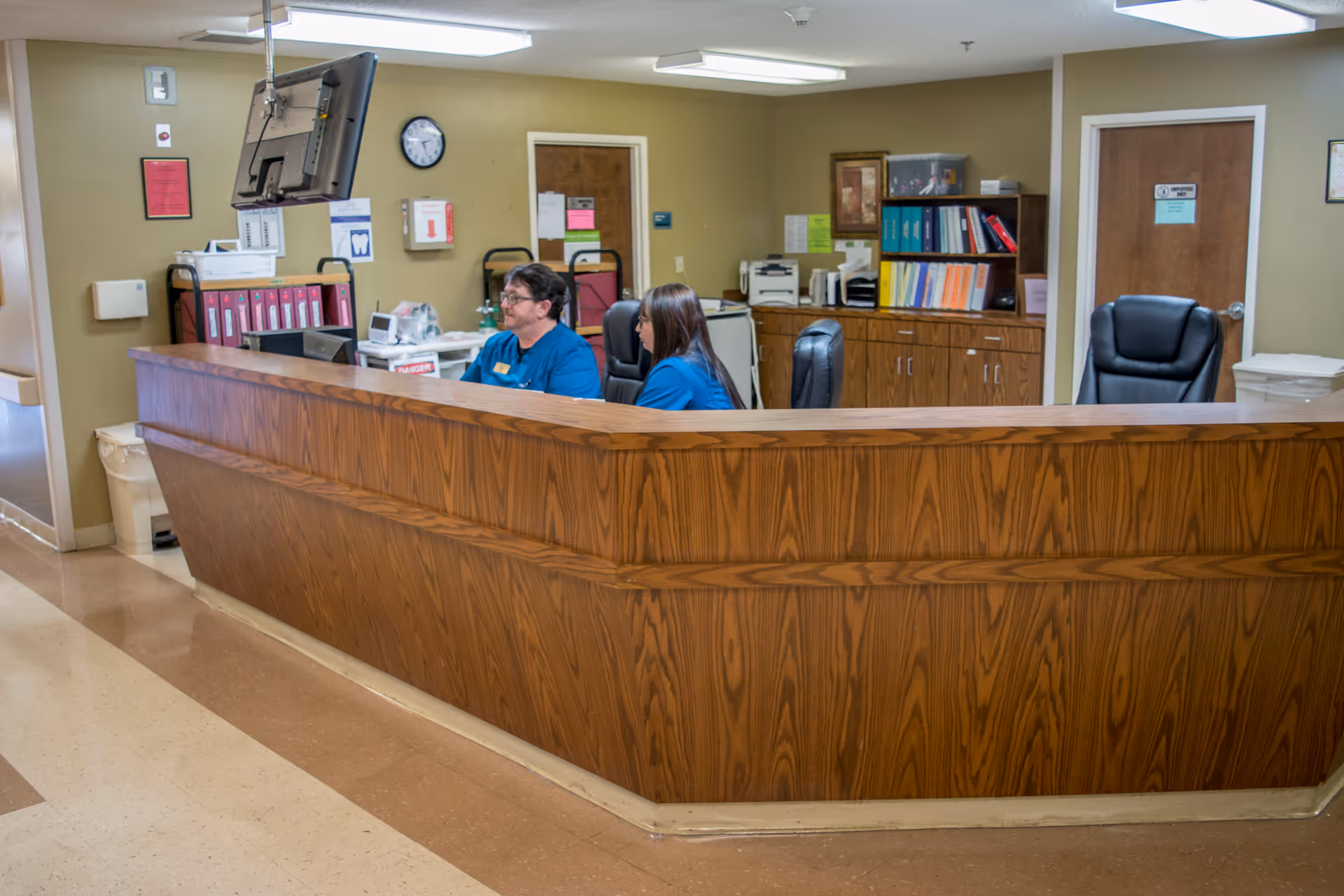 Reception area inside Cleburne County Nursing Home with two staff members seated behind a wooden front desk. The background shows office supplies, filing cabinets, a clock, and two closed doors.