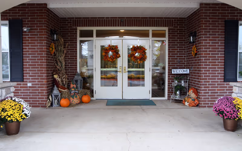 Front entrance of a brick building with double glass doors decorated with fall wreaths, pumpkins, and potted flowers.