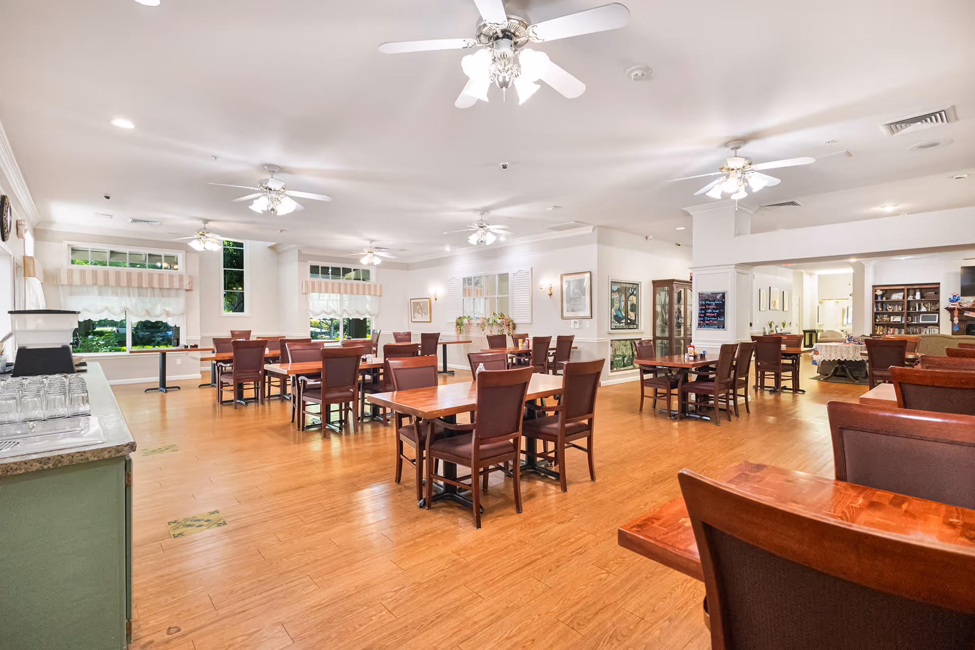 Spacious bright dining room with multiple wooden tables and chairs, ceiling fans, large windows, and hardwood floors.