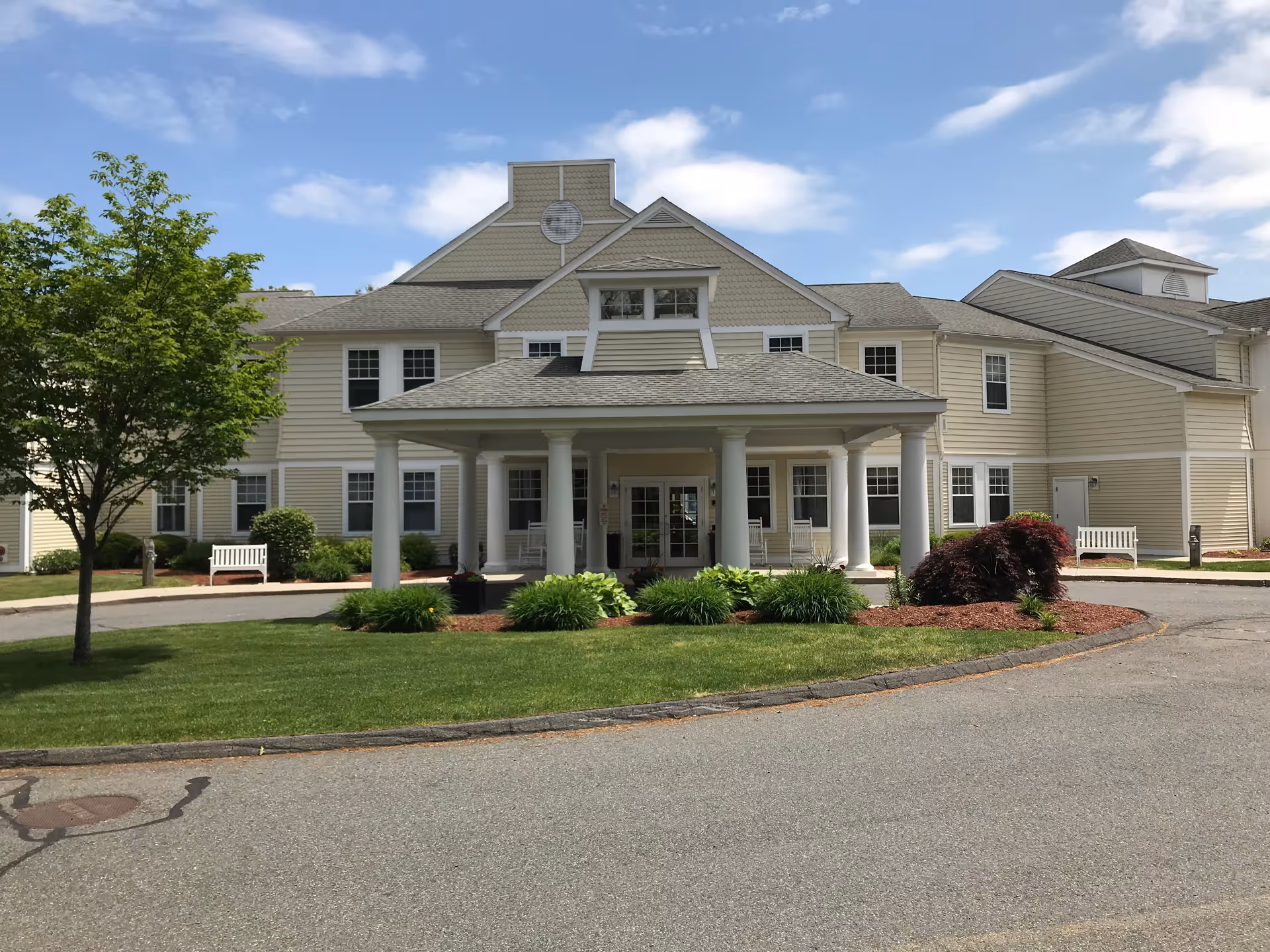 Front entrance of a beige two-story assisted living building with a covered porte-cochère, columns, benches, and landscaped lawn.