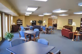 A spacious common area with multiple tables and chairs arranged for seating. The room has beige walls, carpeted floors, and several ceiling lights. There are bookshelves, potted plants, and comfortable armchairs and sofas along the walls, creating a welcoming lounge environment.