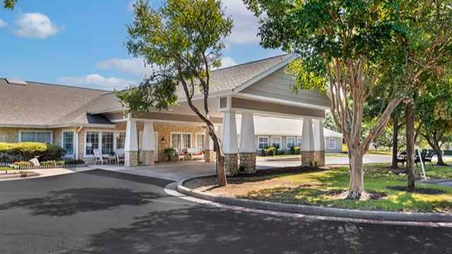 Covered porte-cochere entrance to a senior living building with trees and a curved driveway.