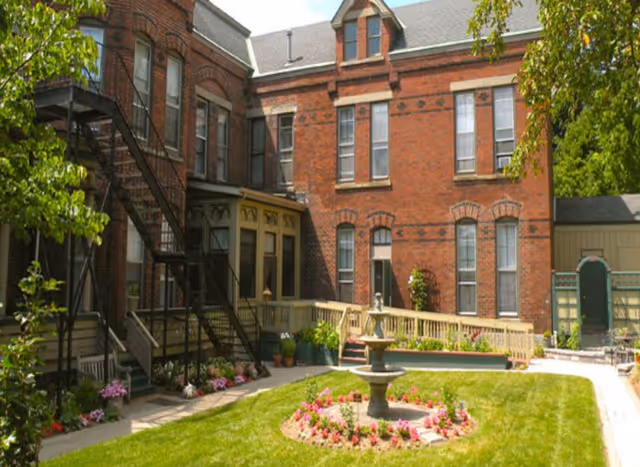 A sunny courtyard with a green lawn and a circular flower bed surrounding a tiered water fountain. The courtyard is enclosed by a red brick building with multiple windows and an external metal staircase. There are flower pots and plants along the walkway and a wooden ramp leading to a door.